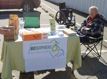 An older woman seated outside in a parking lot next to a REquipment donation day table with banner and brochures and donuts. A wheelchair in the background.