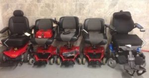 Five power wheelchairs of various types lined up against a wall. 