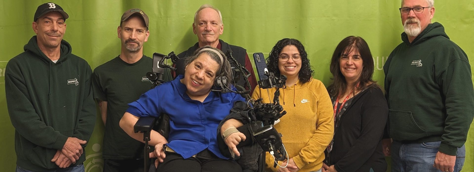 A group of standing smiling adults including a woman in a complex power wheelchair. 