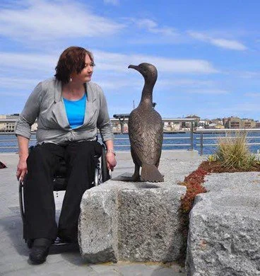Woman in wheelchair staring into the eyes of a bronze duck sculpture with harbor in background. 