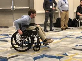 A smiling man racing in a manual wheelchair in a conference hall with onlookers.A smiling man racing in a manual wheelchair in a conference hall with onlookers.