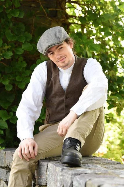 A young man wearing a cap, smiles and poses informally, seated on a stone wall.
