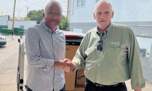 Two men face the camera on a loading dock and shake hands. A van is behind with rear doors open showing an interior packed with boxes.