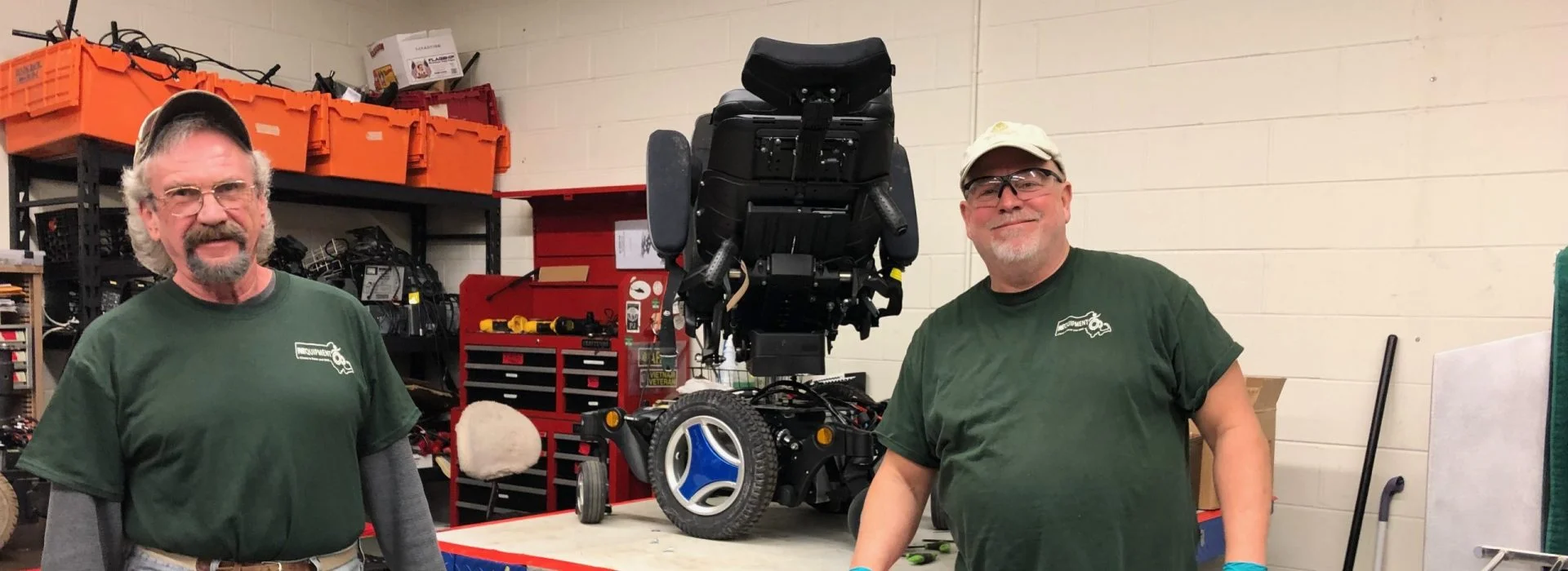 two male staff members smiling with a power wheelchair on a workbench. 